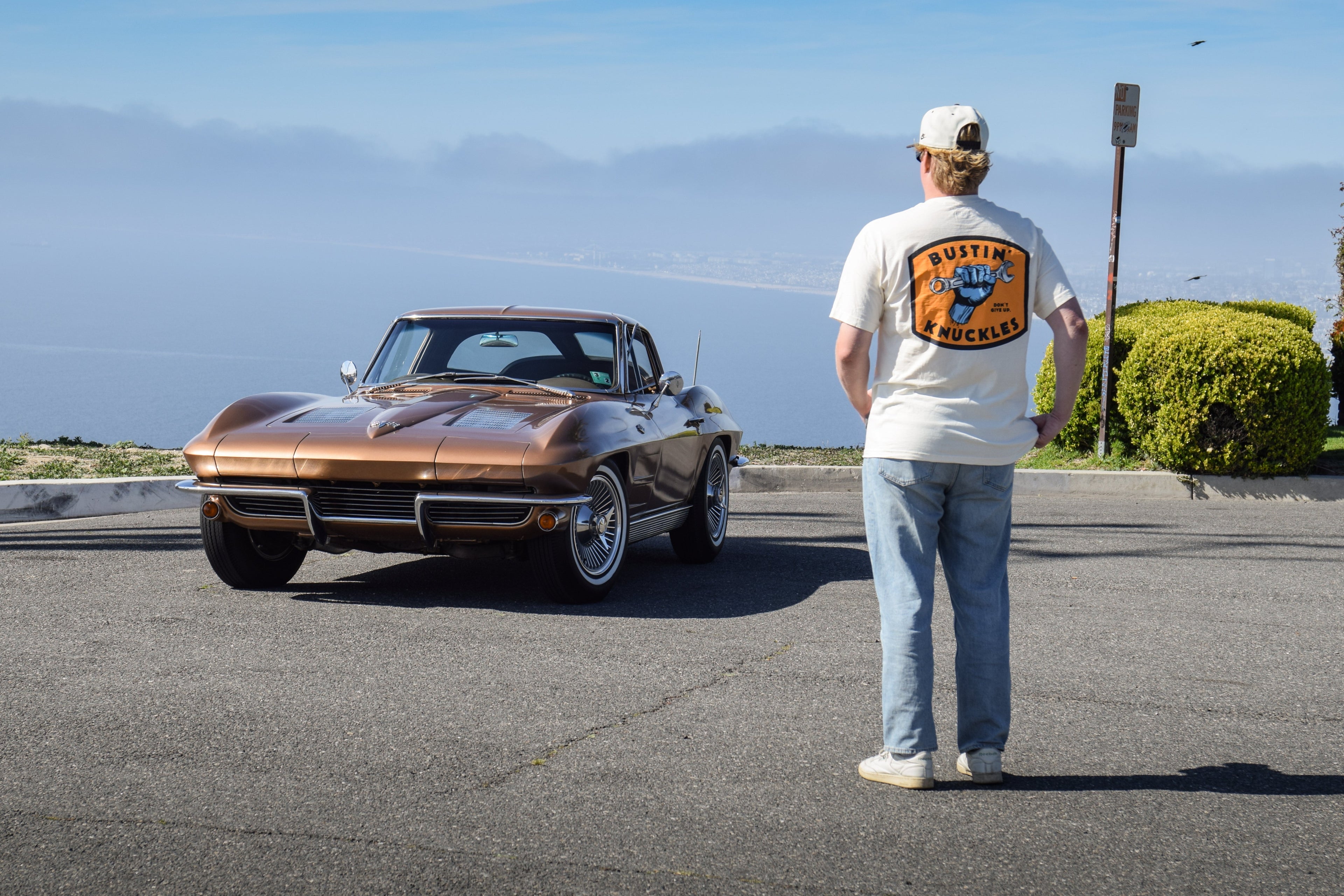 Man wearing cream Stedfast "Bustin' Knuckles" tee and looking at a gorgeous brown 1963 corvette at an ocean overlook. Automotive lifestyle photo